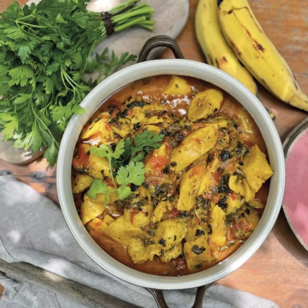 Panela com prato vegano de banana-da-terra e ervas frescas, ao lado de bananas e salsa, na cozinha do Karuh