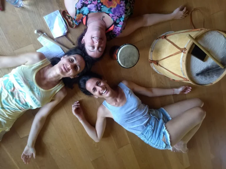 Três mulheres deitadas no chão sorrindo ao lado de tambores